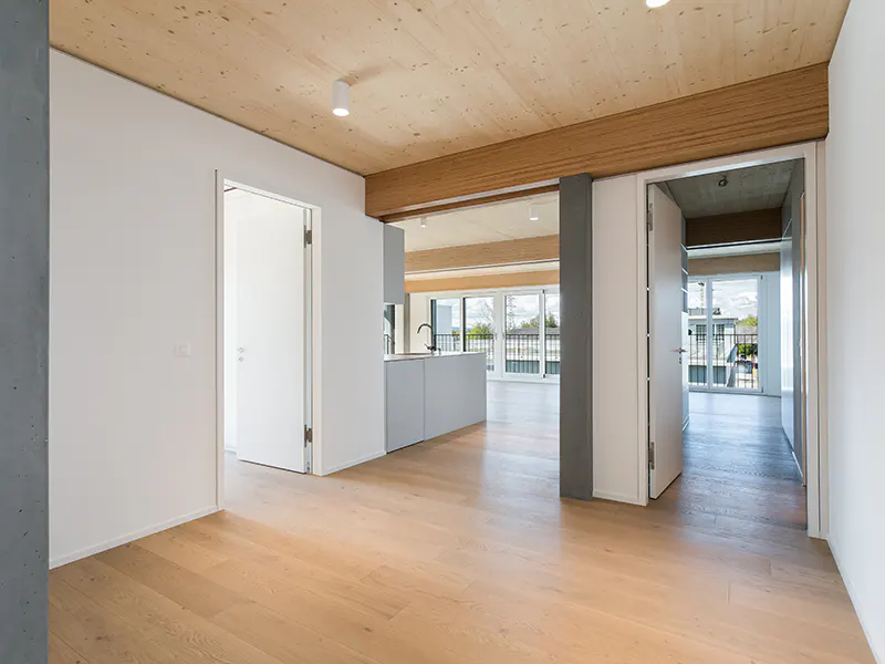 Bright, modern apartment interior with light wood floors, white walls, and a wood ceiling. Open doorways lead to a kitchen and balcony.