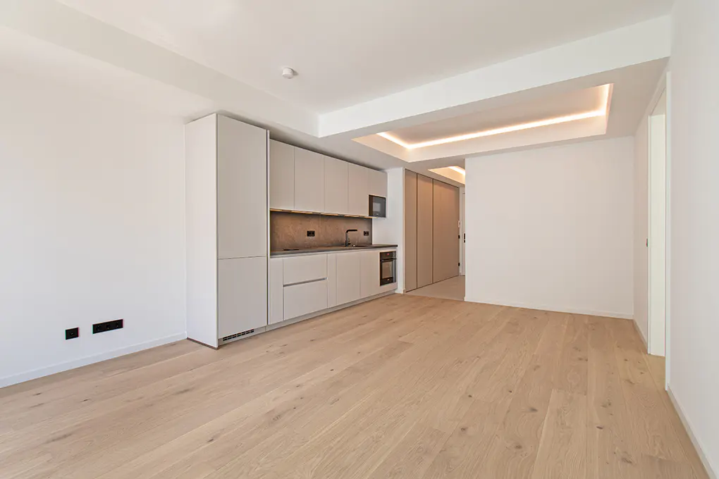 Modern minimalist kitchen with light grey cabinets, wood flooring, and recessed ceiling lighting in an open-plan room.