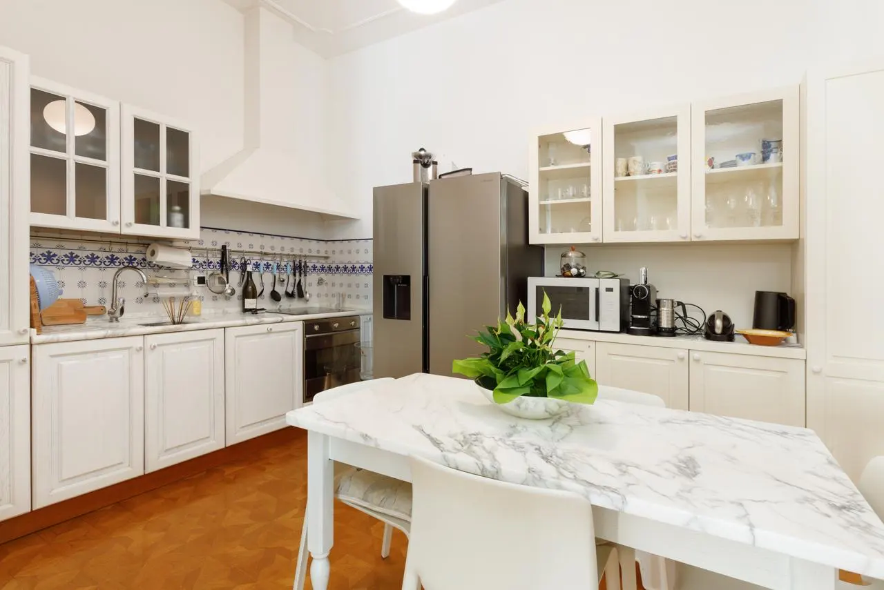 Bright kitchen with white cabinets, marble countertops, and stainless steel fridge. A marble-topped table with a plant sits in the foreground.