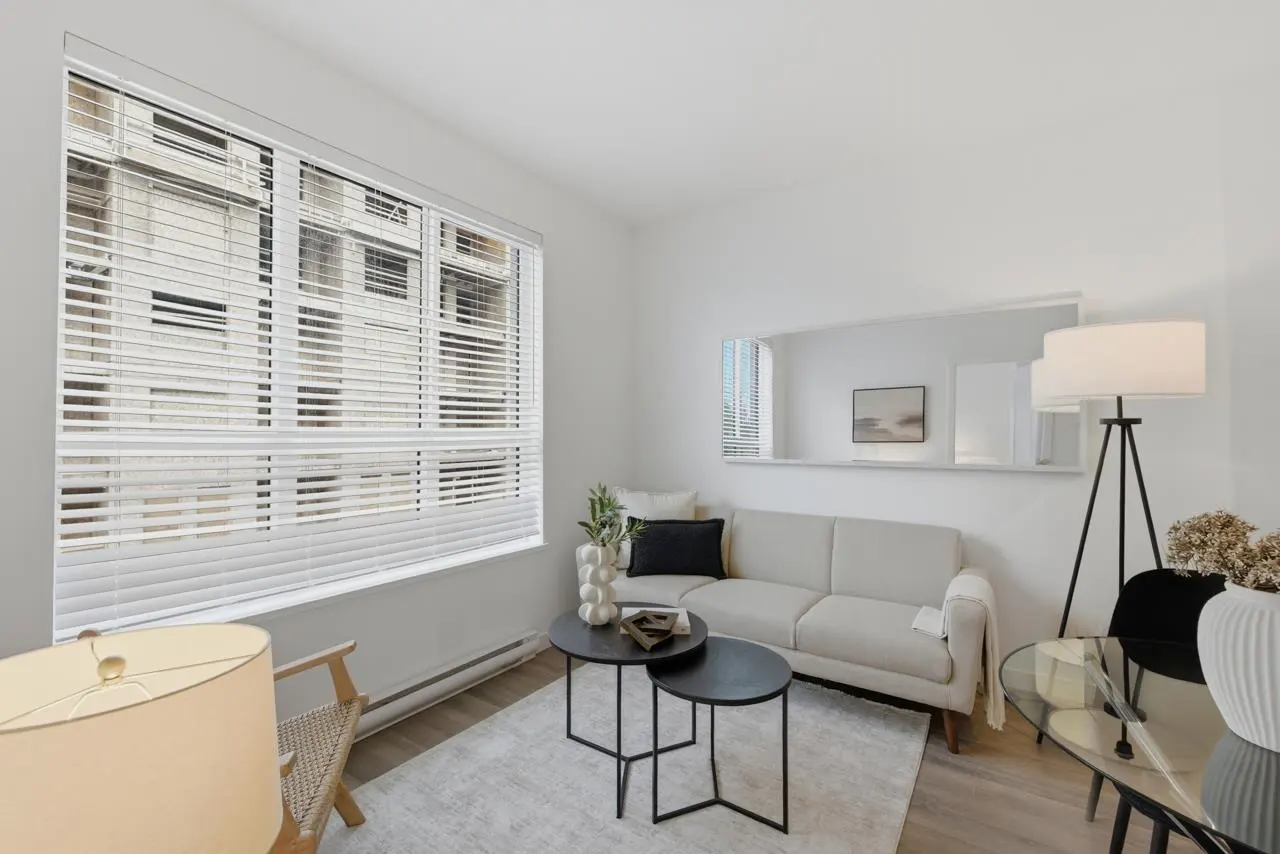 Bright, modern living room with a beige sofa, black nesting tables, and a large window with white blinds. A floor lamp and mirror add depth.