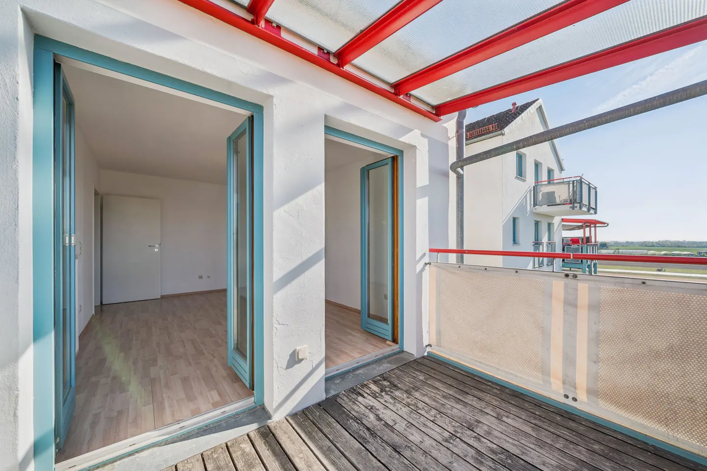 Balcony with wooden floor, white walls, and blue-framed doors leading to a room with light wood floors. Red metal roof structure overhead.