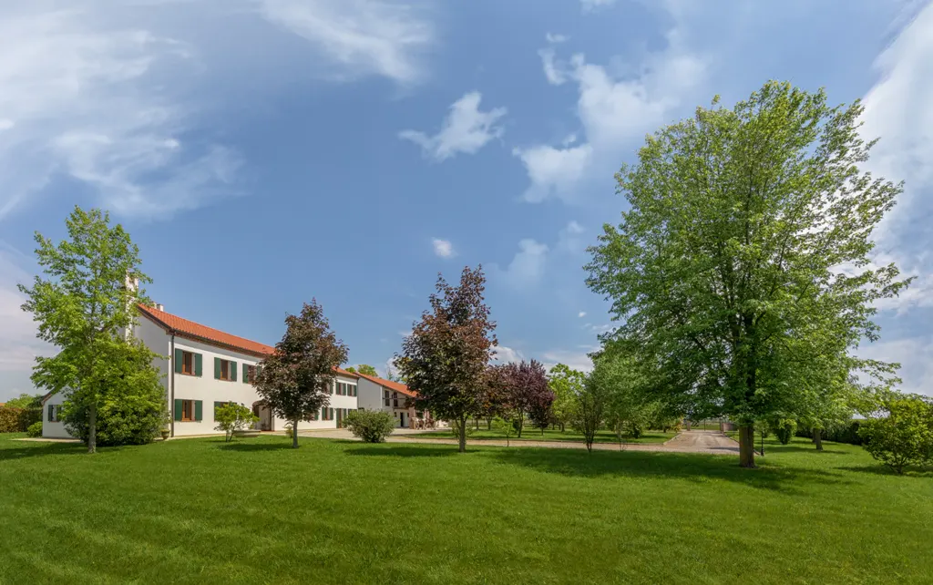A white building with a red roof and green shutters sits on a green lawn under a blue sky with scattered clouds. Trees surround the building.