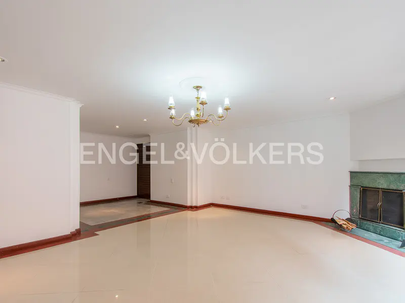 Empty living room with white walls, tile floors, and a green marble fireplace. A chandelier hangs from the ceiling. Engel & Völkers logo is visible.