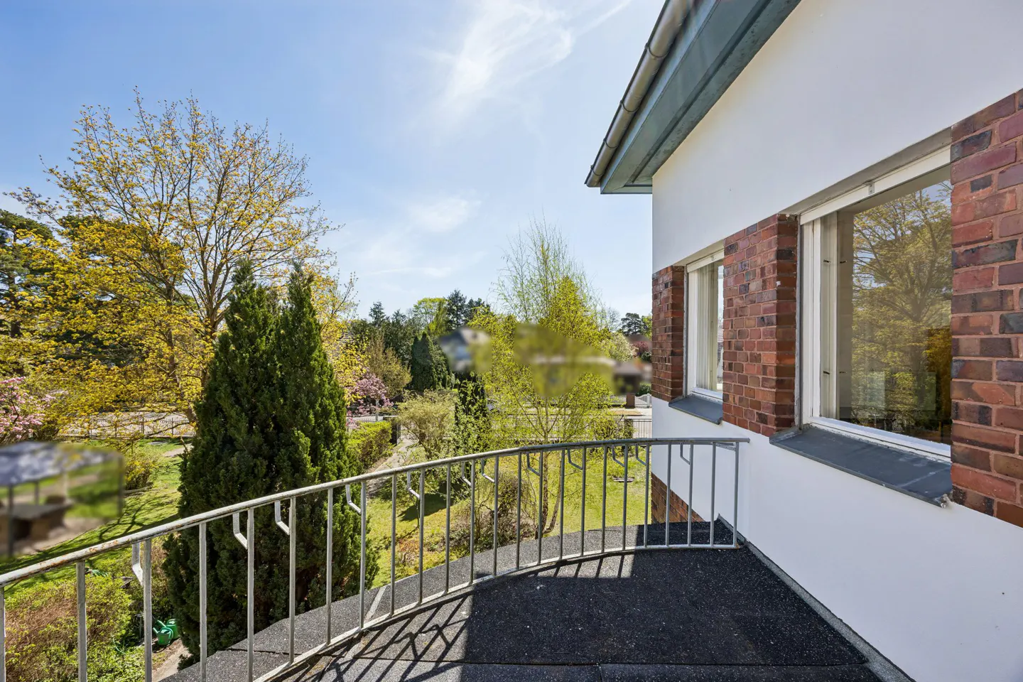 View from a balcony with a metal railing overlooking a green garden with trees and a white building with brick accents.