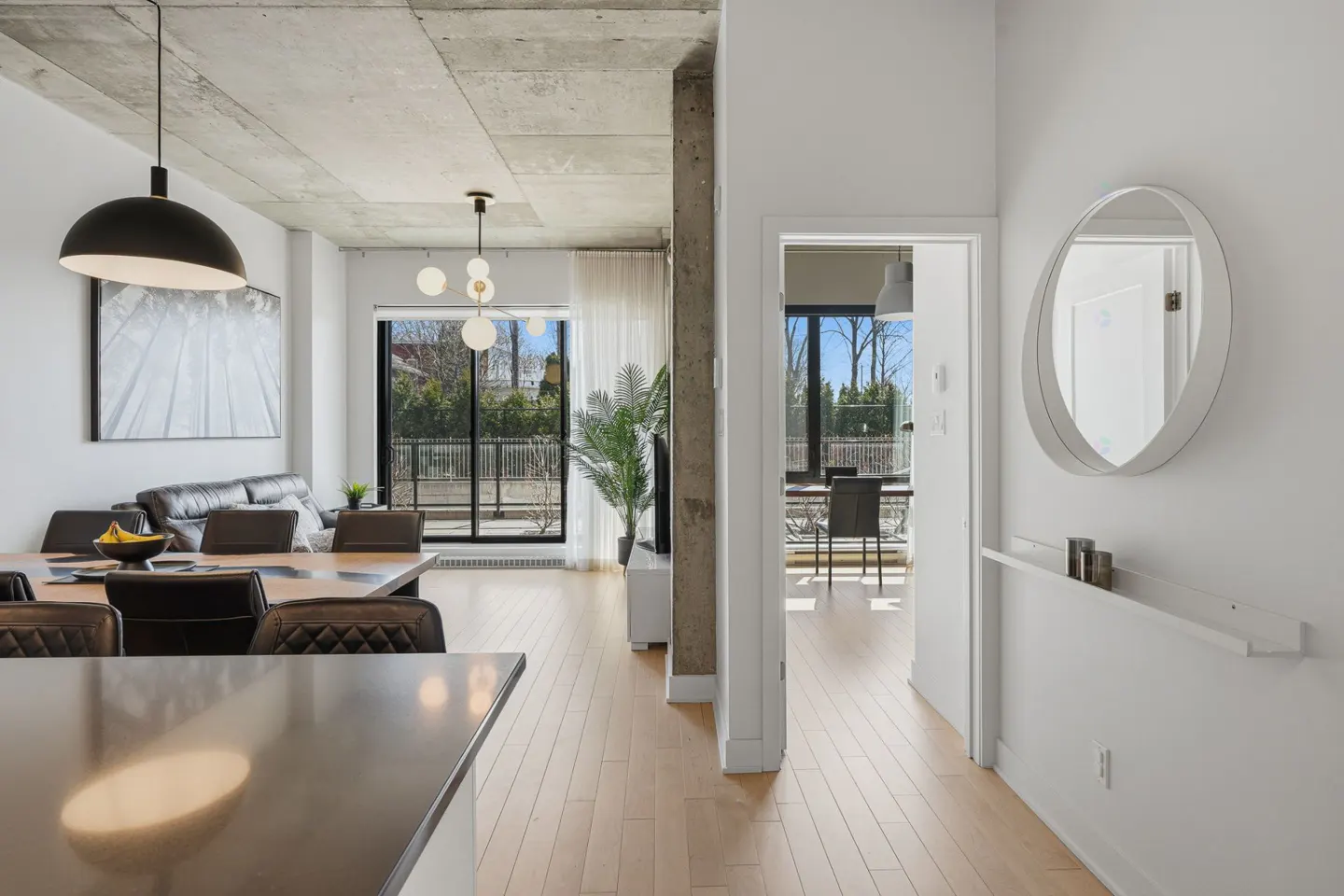 Modern apartment interior with concrete ceiling, wood floors, and white walls. A dining table, sofa, and balcony are visible.