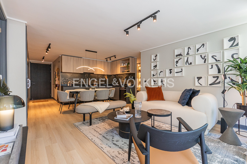 A modern apartment interior with a white sofa, dining table, and kitchen. Artwork adorns the wall. The color palette is neutral with pops of orange and black.