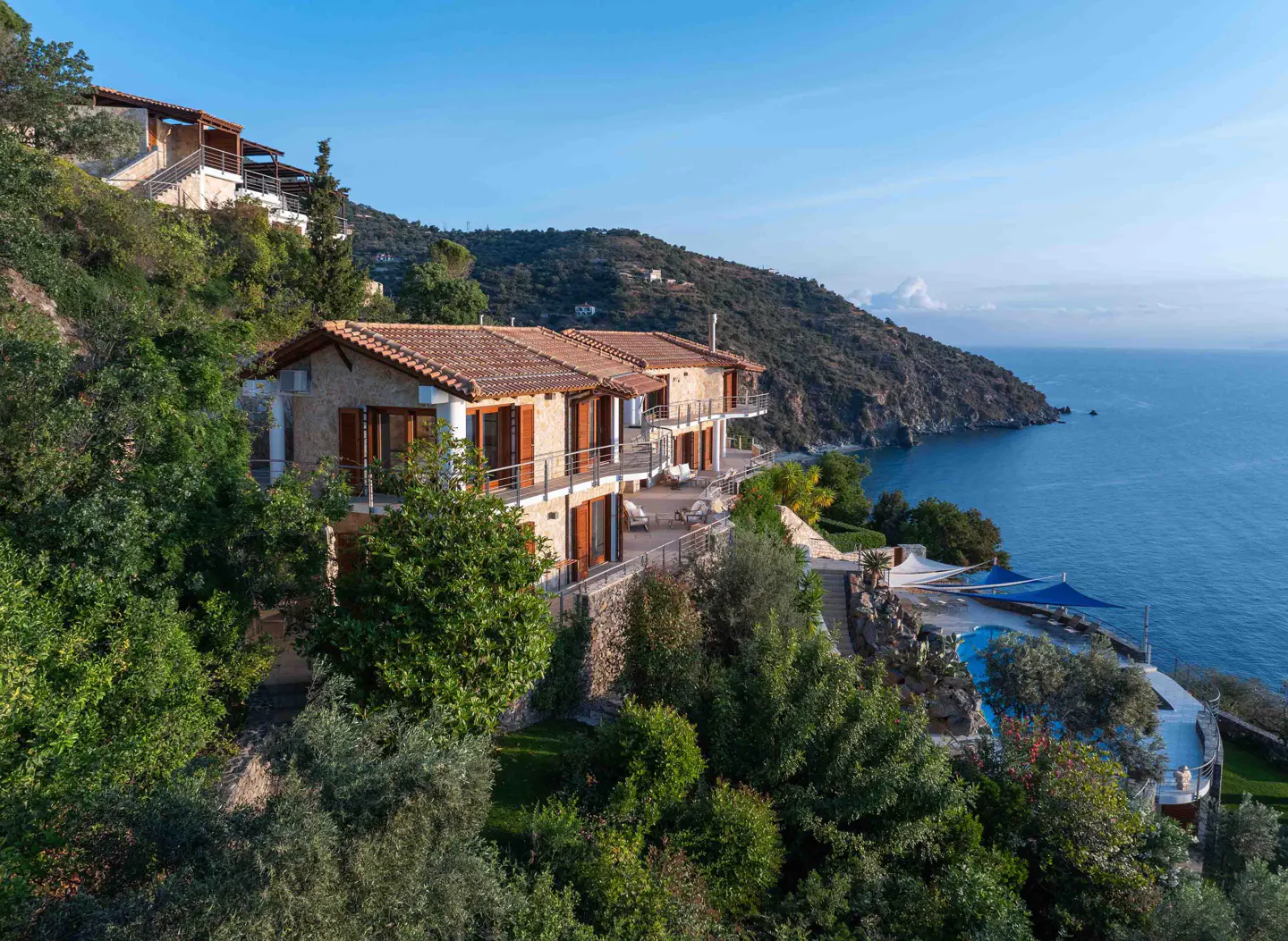 Exterior view of a stone house with a red tile roof on a green hillside overlooking the blue ocean.