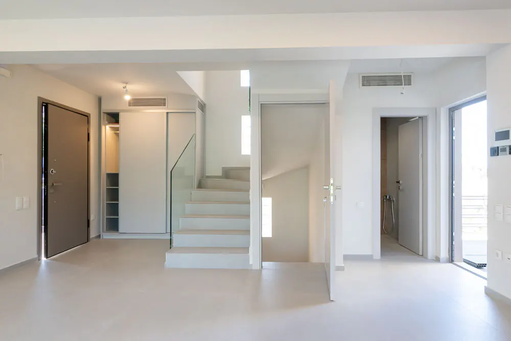 Modern minimalist interior with white walls, light flooring, a staircase with glass railing, and several doorways.