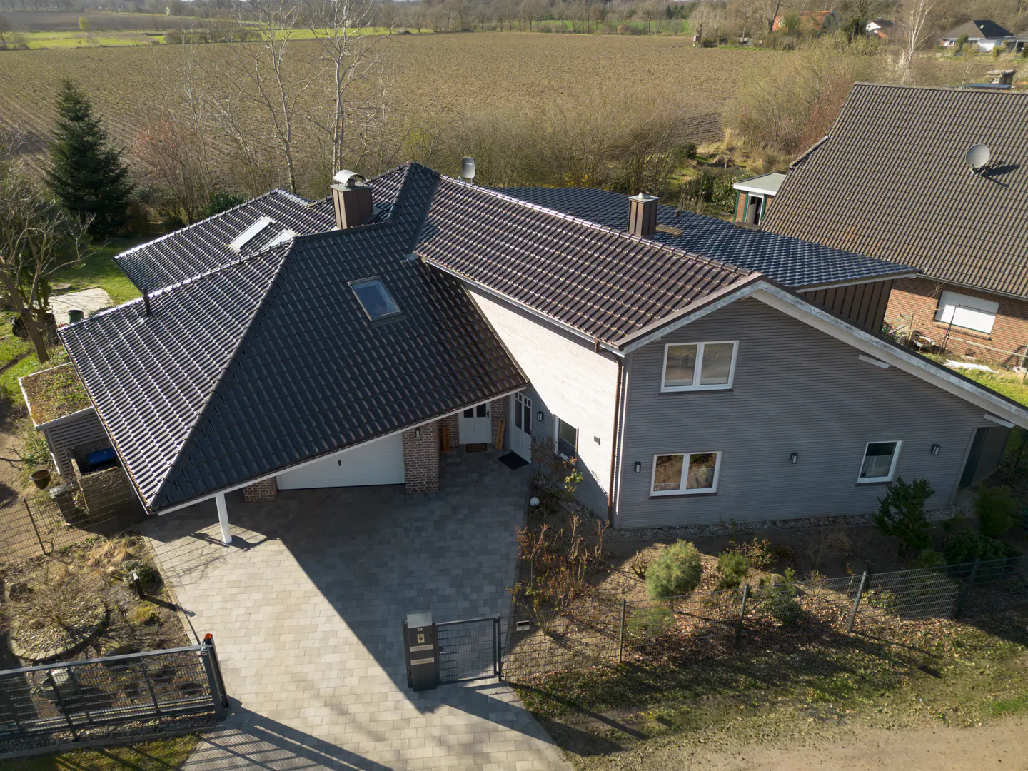 Aerial view of a gray, two-story house with a dark gray tiled roof, a paved driveway, and a gated entrance.
