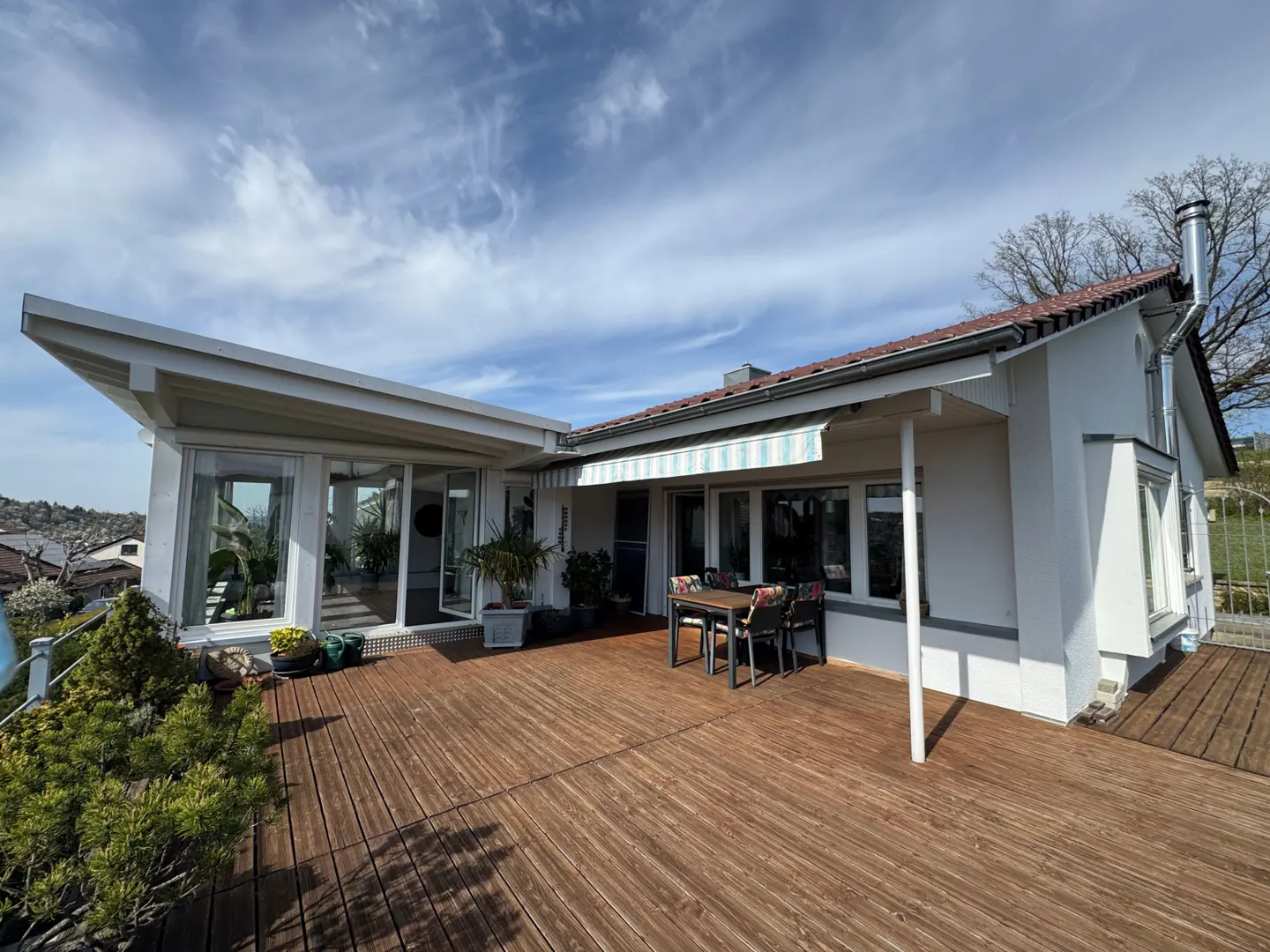 Exterior view of a white house with a wooden deck, table, and chairs under a blue and white awning.