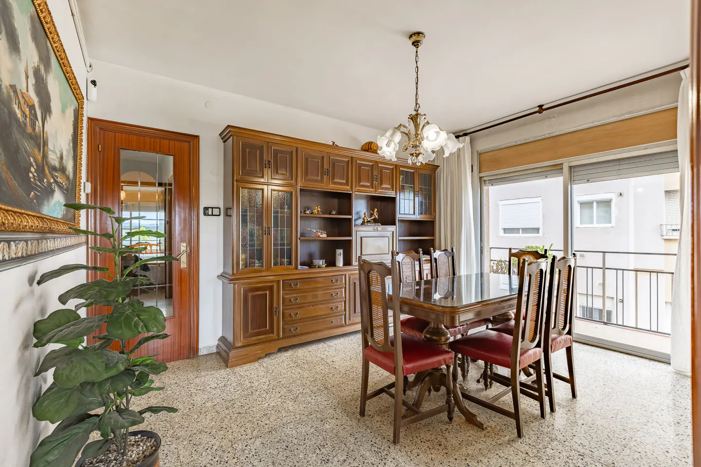 A dining room with a wooden table, red chairs, a large wooden cabinet, and a balcony with a view.
