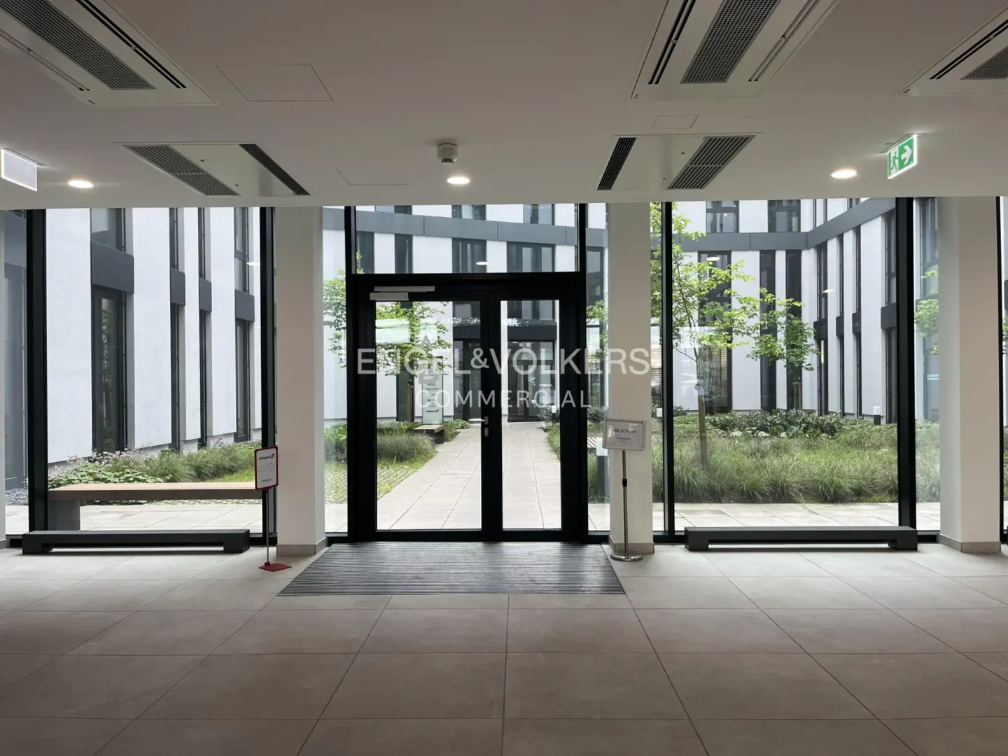 A bright lobby with large windows and glass doors leading to a courtyard. The Engel & Volkers logo is on the glass.