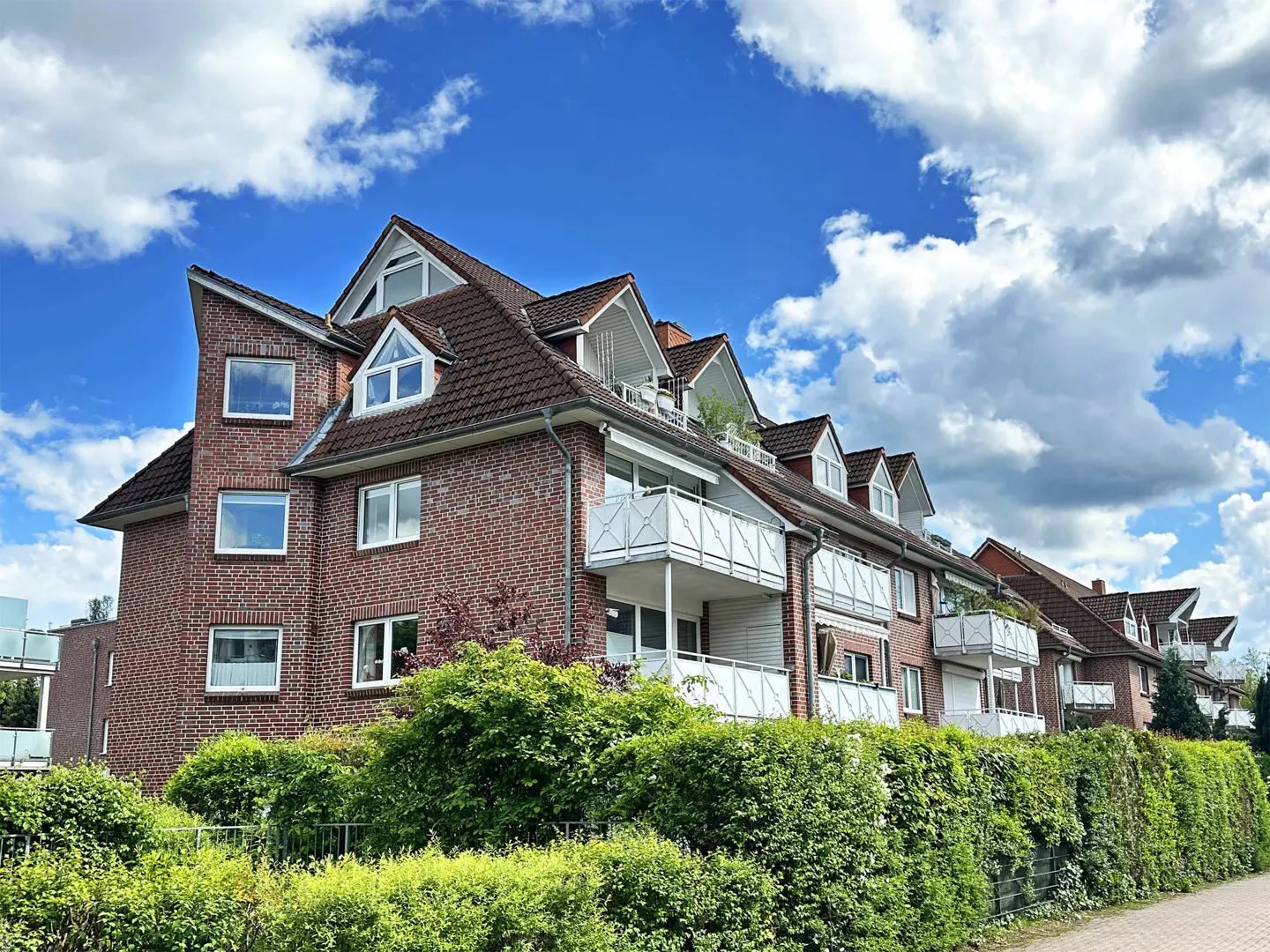 Brick apartment building with white balconies and dormer windows under a blue sky with fluffy white clouds. Green bushes line the front.