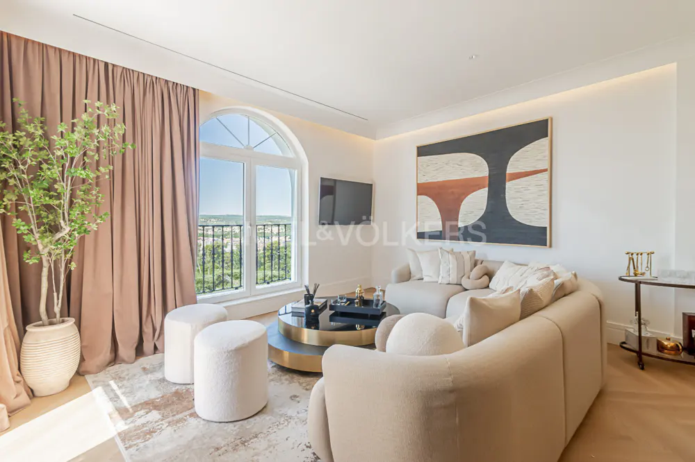 Bright living room with beige sofa, round coffee table, and arched window with a view. Artwork hangs on the white wall.