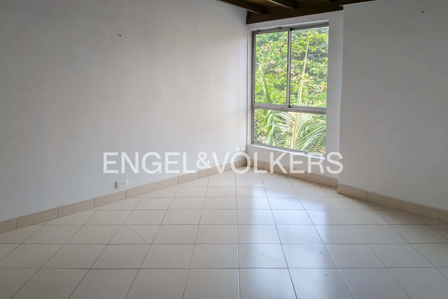 Empty room with white walls, tile floor, and a large window showing green trees. Engel & Völkers logo is visible on the wall.