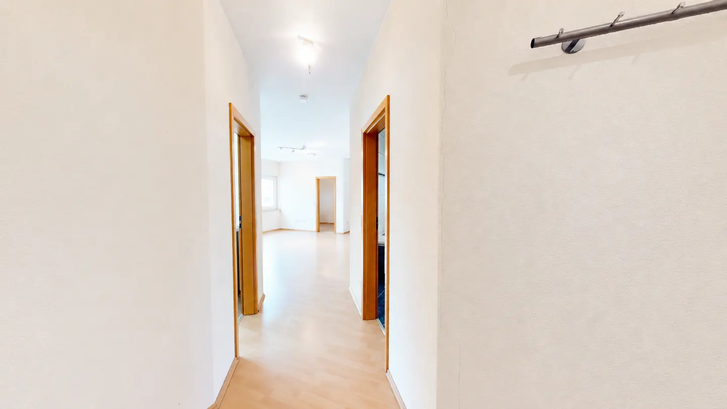 Hallway with light wood floors and white walls. Two open wood doors lead to rooms with windows. A metal coat rack is on the right wall.