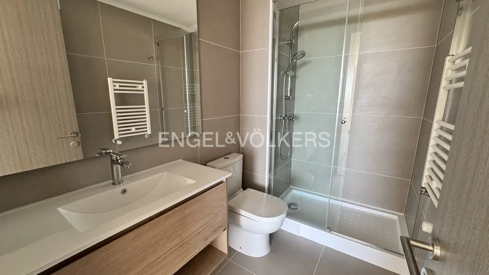 Bathroom with a white sink, wood vanity, toilet, and glass shower. Gray tiled walls and a white towel rack are visible.