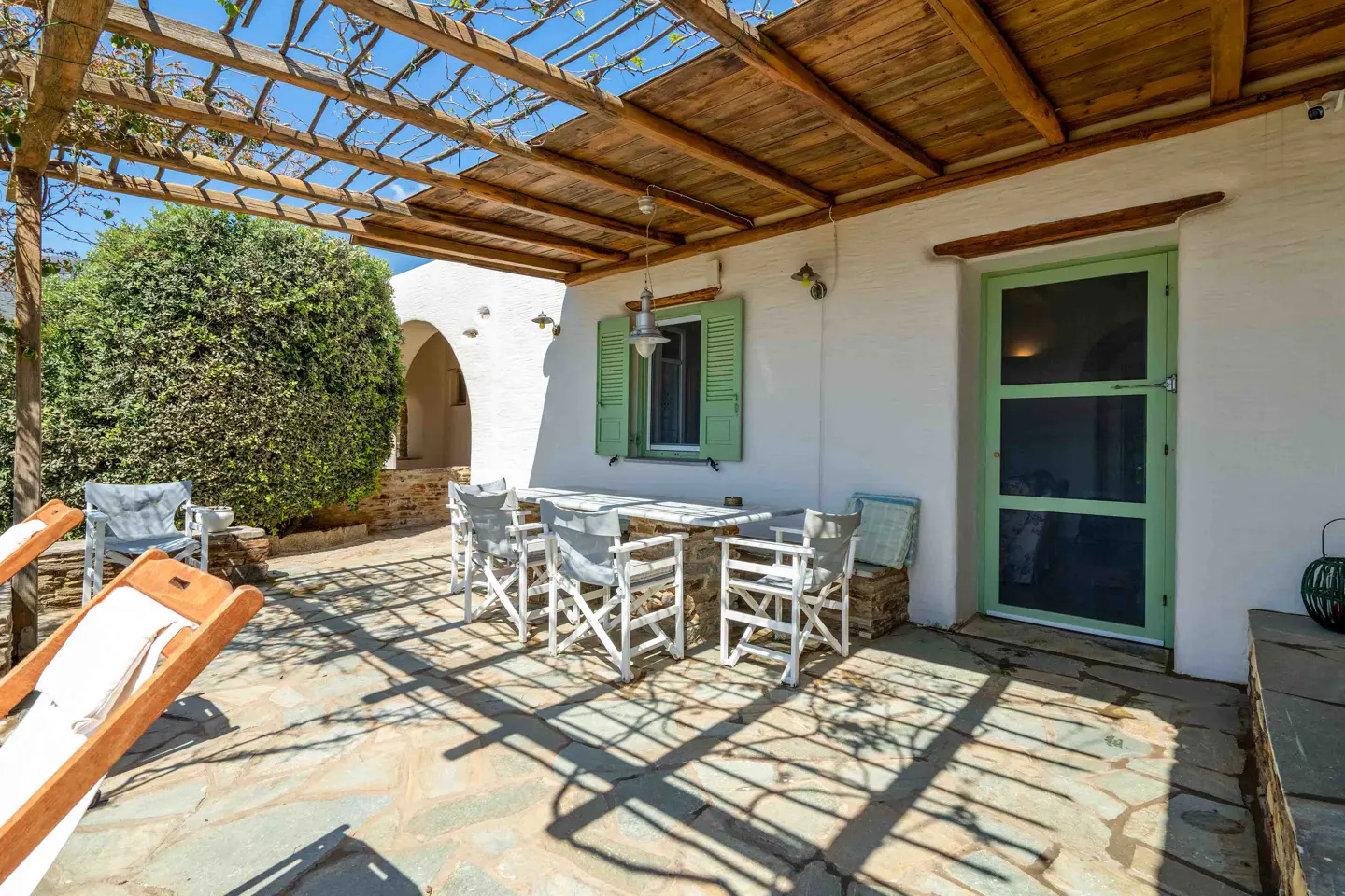 Outdoor patio with a wooden pergola, white walls, and green door and shutters. A table and chairs sit on a stone patio.