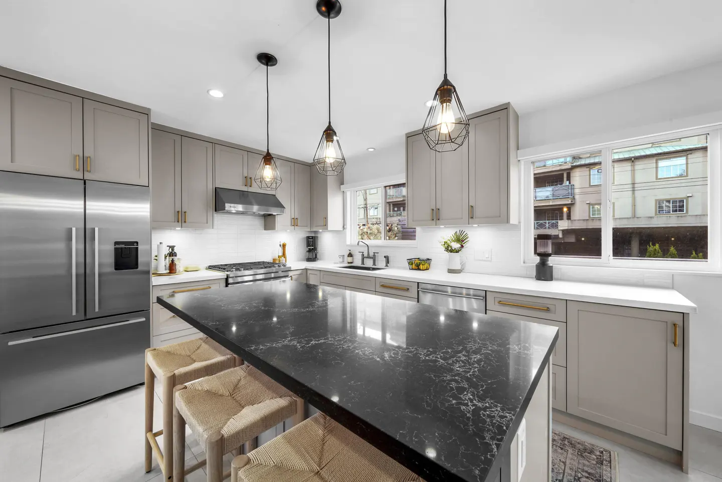 Bright kitchen with gray cabinets, stainless steel appliances, and a black marble island with three woven stools. Three pendant lights hang above.
