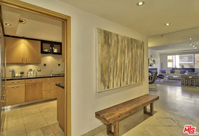 Hallway with a wooden bench under a large abstract painting. A kitchen is visible through a doorway. Living room in the background.