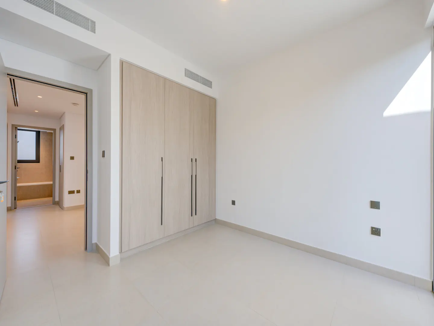 Bright, empty bedroom with white walls, light tile floor, and a large, light wood closet. A doorway leads to another room.