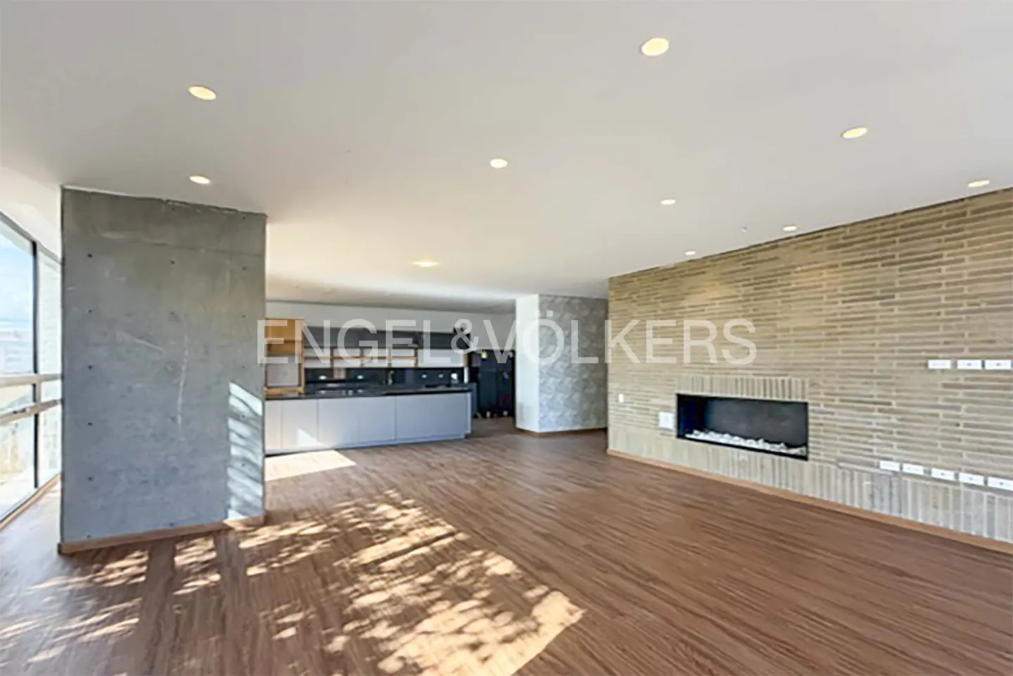 Bright, open-concept living space with wood floors, brick fireplace, and modern kitchen. Large windows offer natural light. Engel & Völkers logo visible.