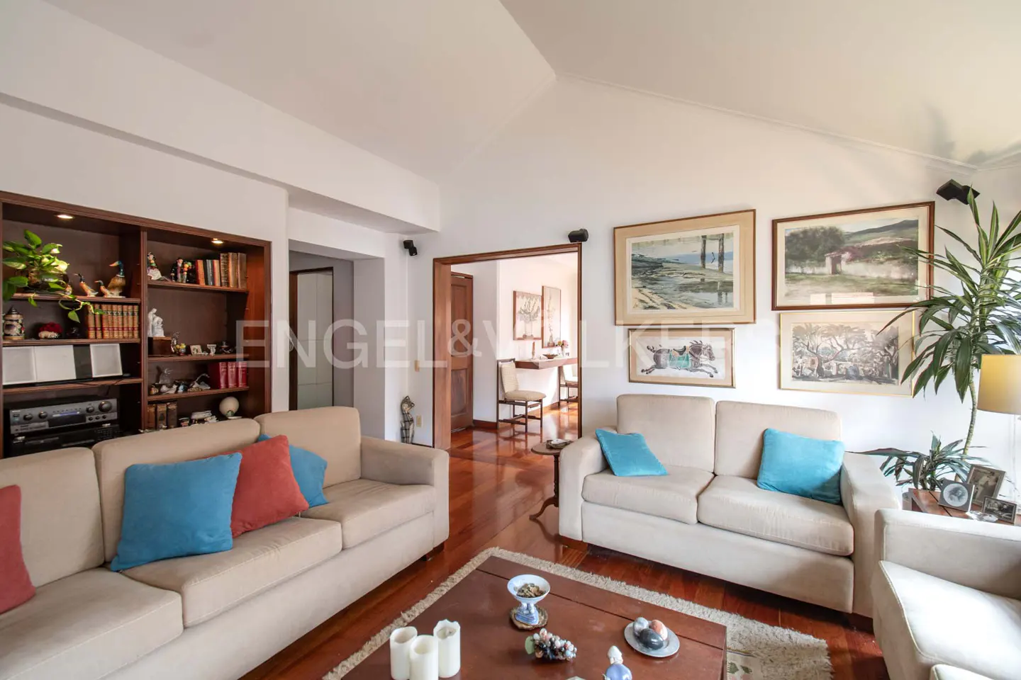 Living room with hardwood floors, beige sofas with blue and red pillows, and a dark wood bookshelf. Artwork hangs on the walls.