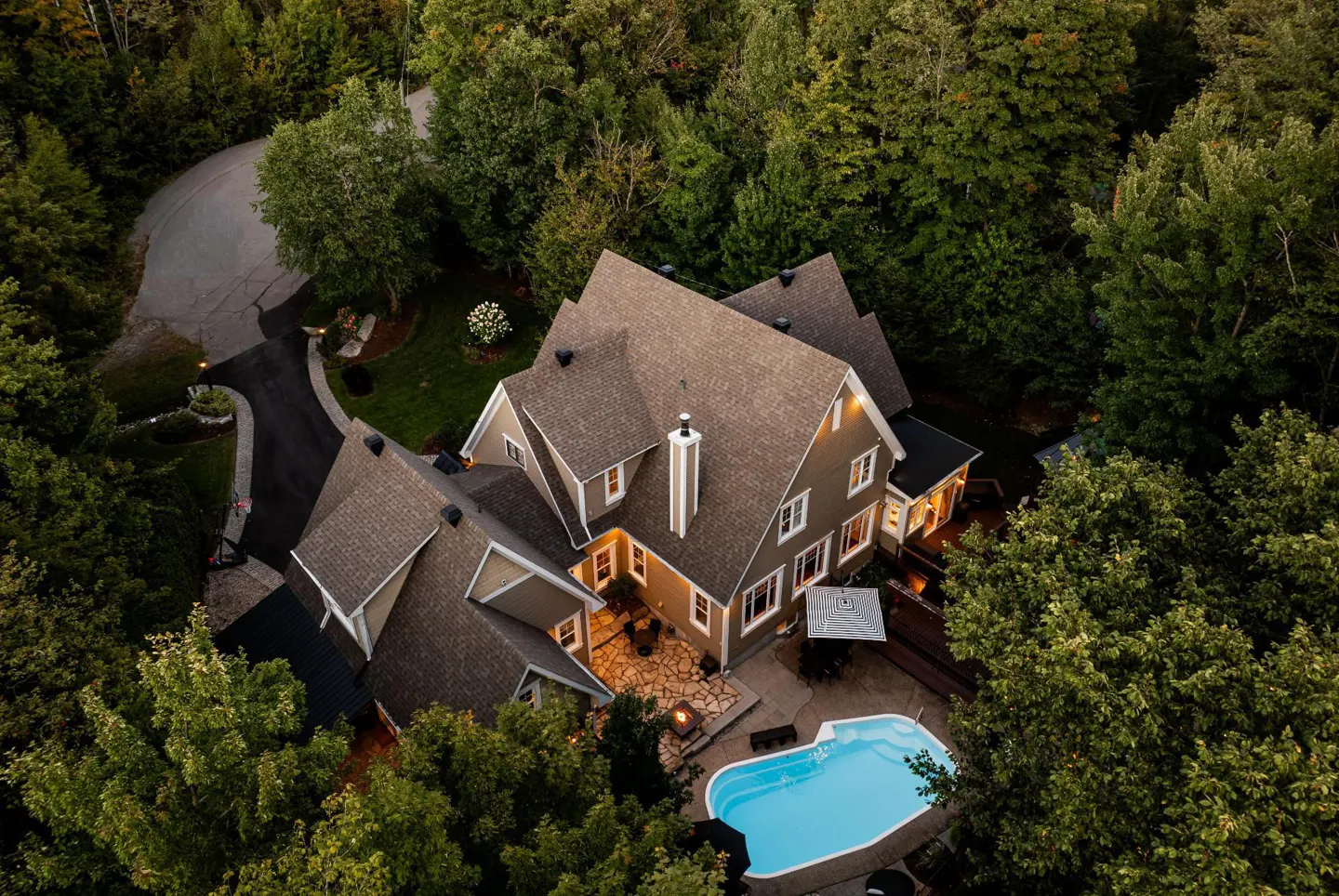 Aerial view of a tan house with a stone patio, swimming pool, and driveway, surrounded by dense green forest.