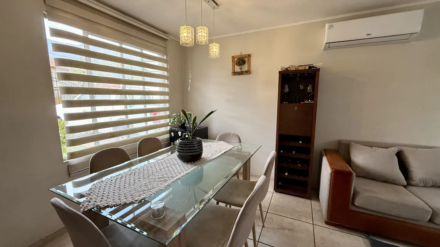 A dining room with a glass table, six chairs, and a brown wooden wine rack. A beige sofa is on the right. The window has horizontal blinds.