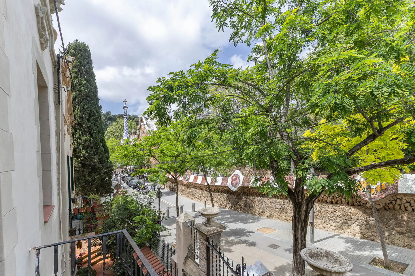 View from a balcony overlooking Park Güell in Barcelona, Spain. Green trees line the street with a mosaic wall and the park's architecture in the background.