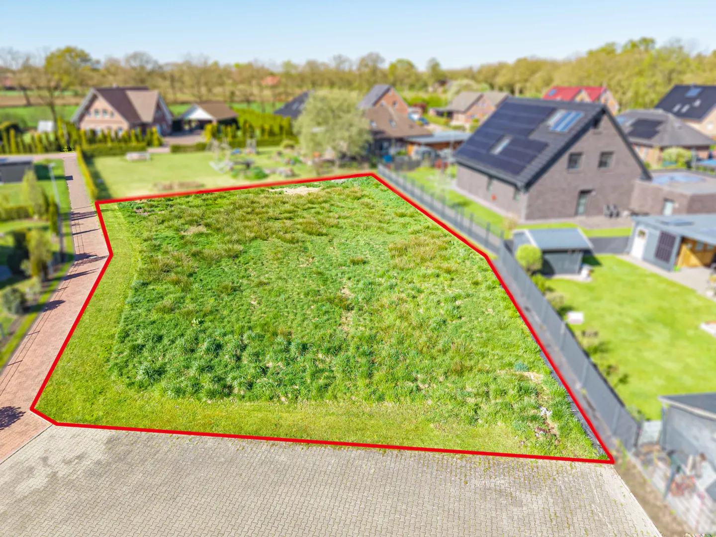 Aerial view of a grassy vacant lot outlined in red, surrounded by houses and trees in a residential neighborhood.