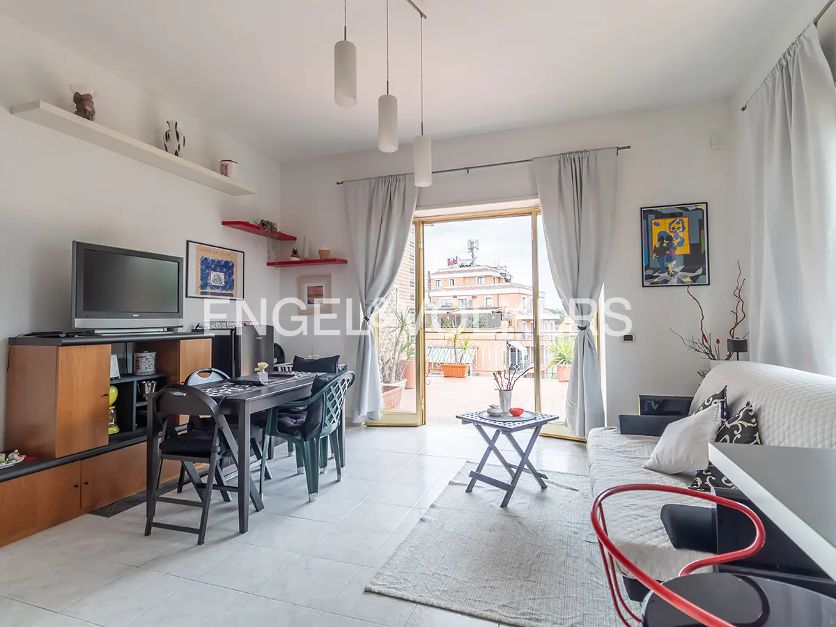 Bright living room with a dining table, TV, and sofa. Balcony doors open to a rooftop terrace with city views. White walls and gray curtains.