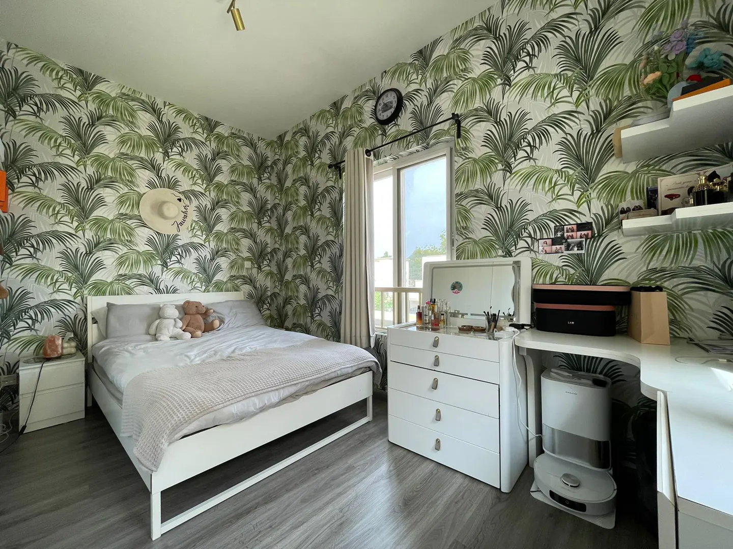 A bedroom with palm tree wallpaper, a white bed with gray linens, and a white vanity with makeup.