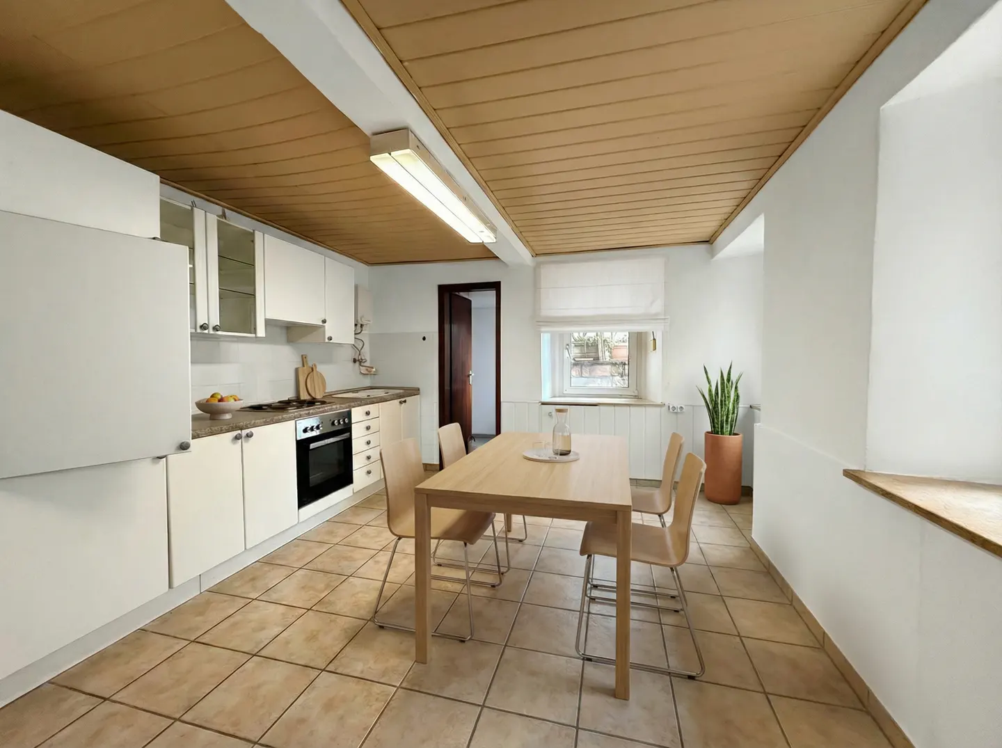 Bright kitchen with white cabinets, a wooden table with four chairs, and a brown tiled floor. A snake plant sits near a window.
