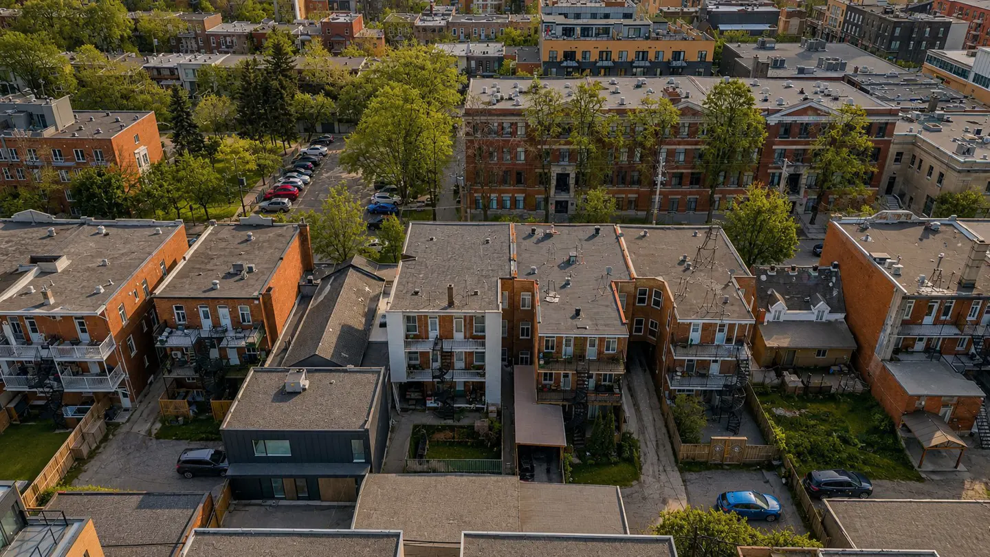 Aerial view of a dense urban neighborhood with brick apartment buildings and green trees. Cars are parked on the street.