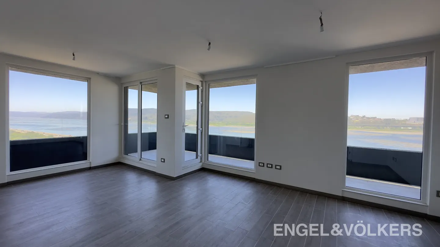 Unfurnished room with wood floors, white walls, and large windows overlooking a lake and green hills. Balcony access via sliding glass doors.
