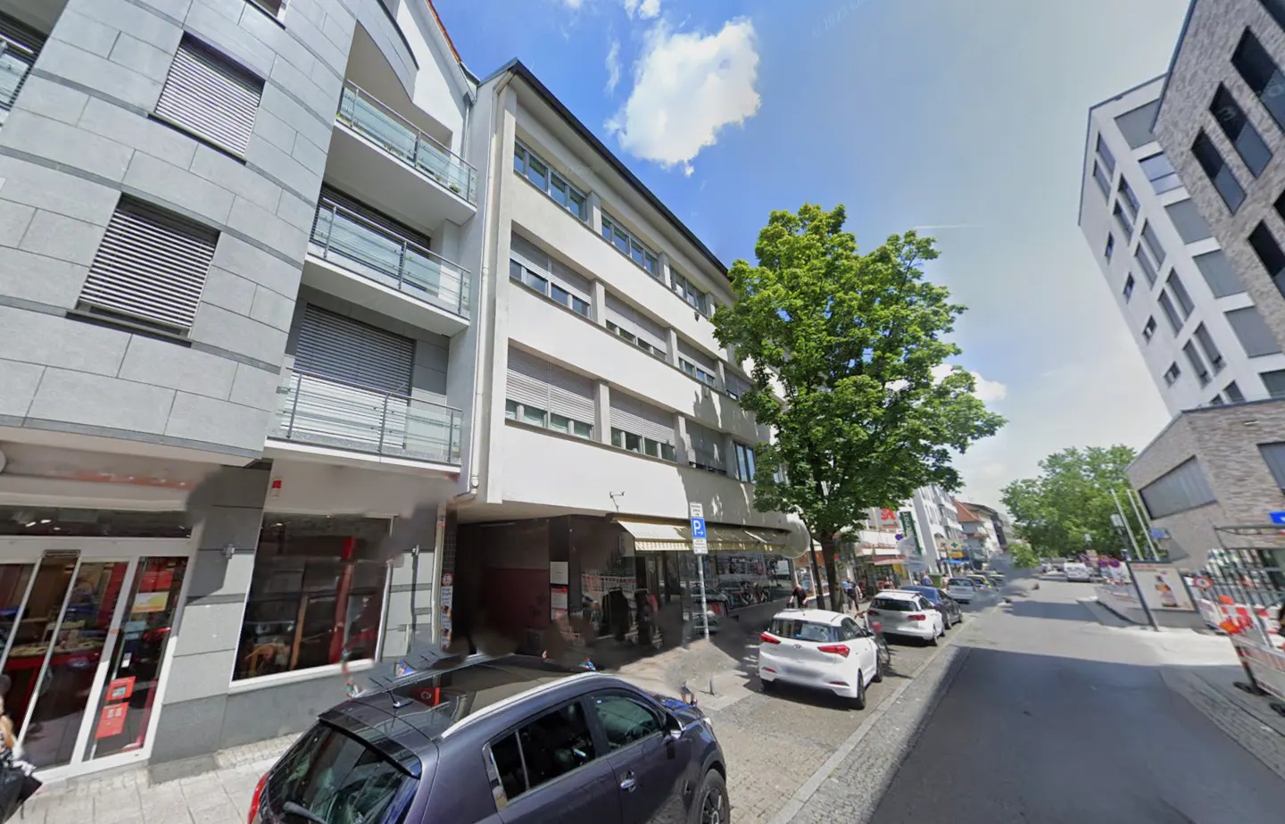 Street view of multi-story buildings with shops and parked cars on a sunny day. A large green tree is in the center.