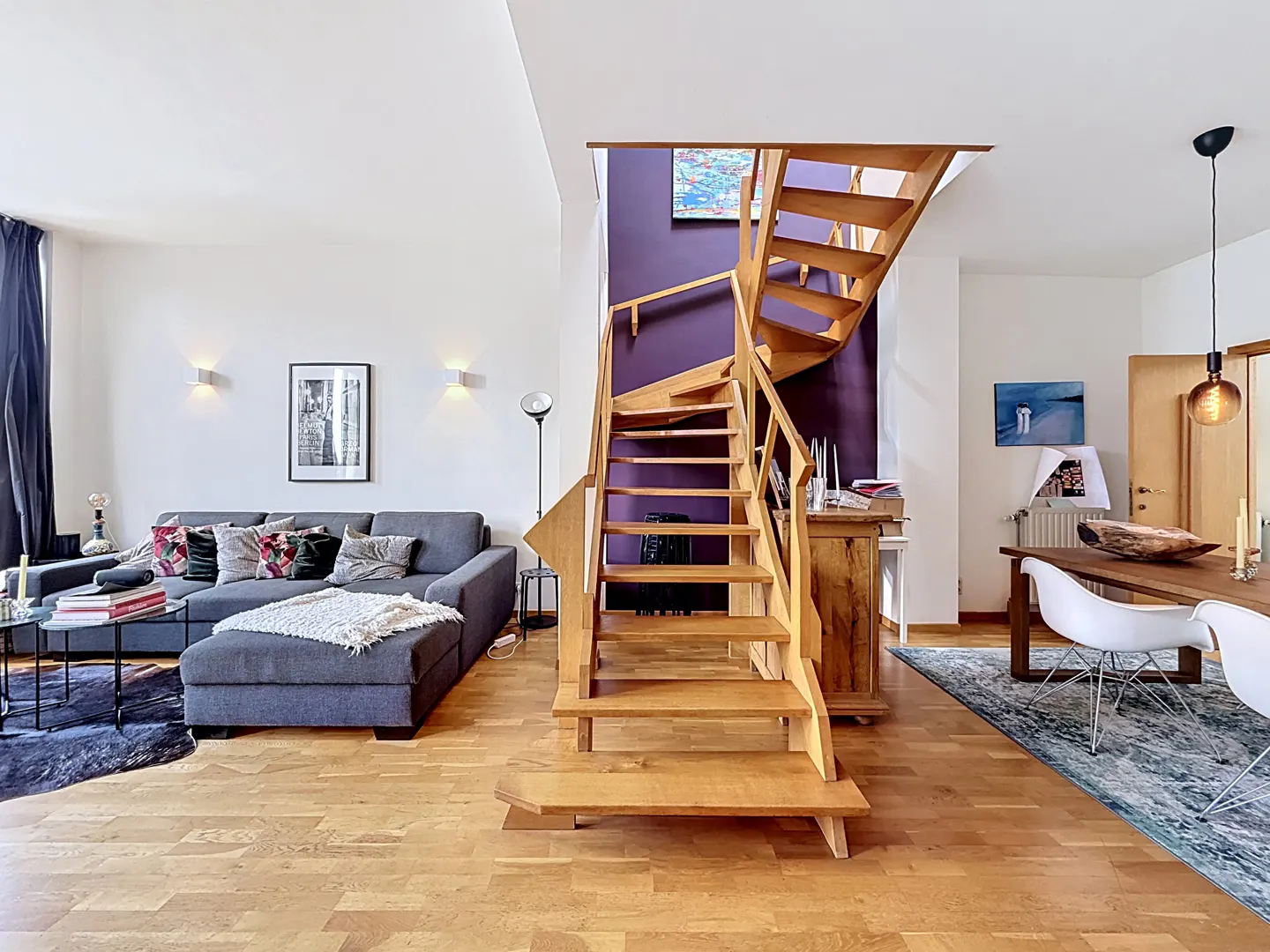 Modern living room with wooden staircase, grey sectional sofa, and dining area with white chairs on hardwood floors.
