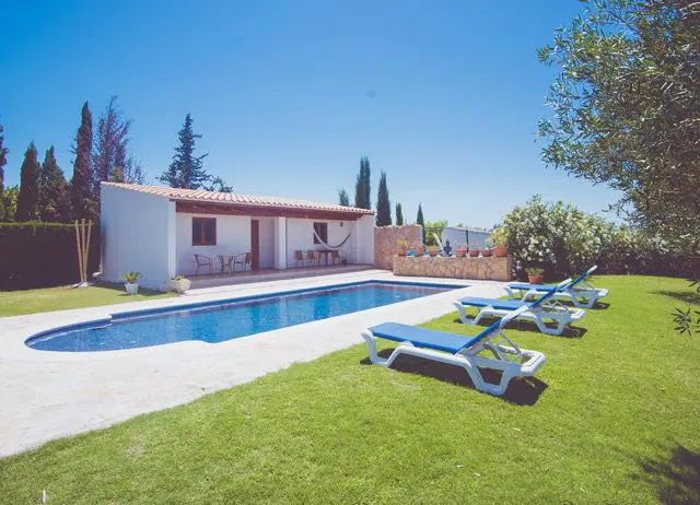A bright, sunny backyard with a pool and lounge chairs. The pool is blue and surrounded by a concrete patio and green grass. A white house with a red tile roof is in the background.