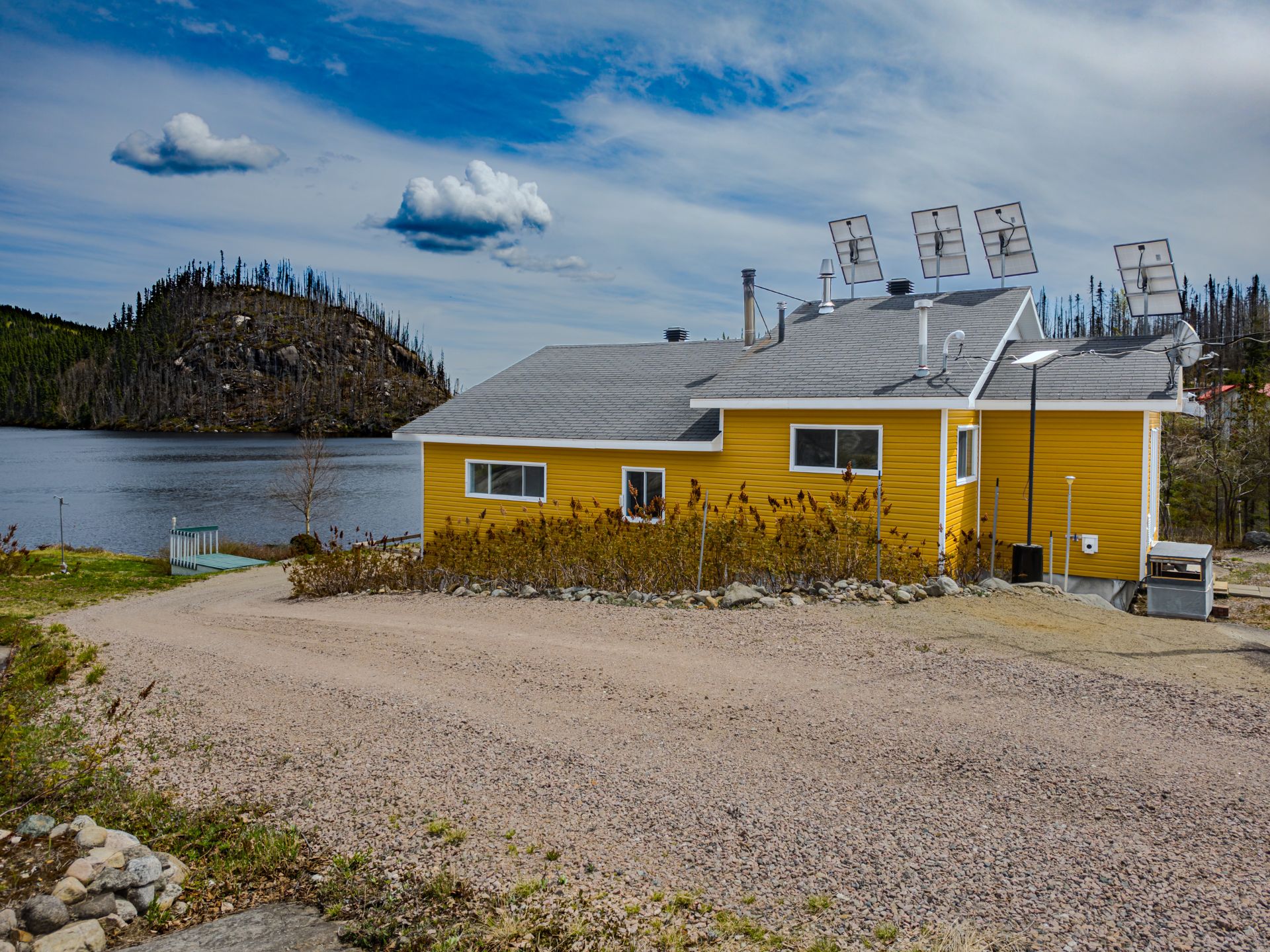 Bright yellow house with solar panels on the roof, situated on a gravel driveway next to a lake and a rocky, forested hill. [AI-generated text]