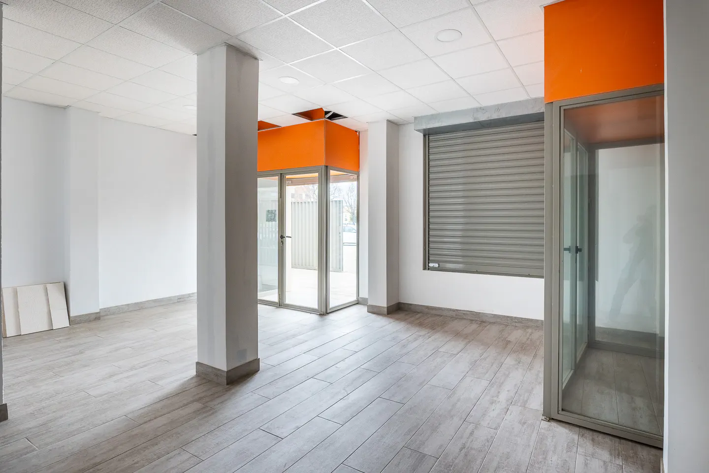 Empty commercial space with gray wood-look floors, white walls, and a white grid ceiling. Orange accents highlight the glass-enclosed entrance and window with a closed gray shutter.