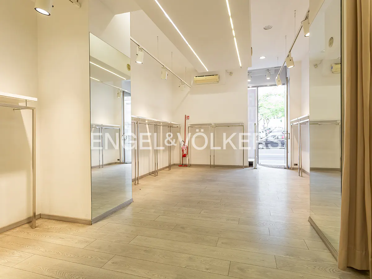 Bright retail space with light wood floors, white walls, and metal clothing racks. Large mirrors reflect the interior. A red fire extinguisher is visible.
