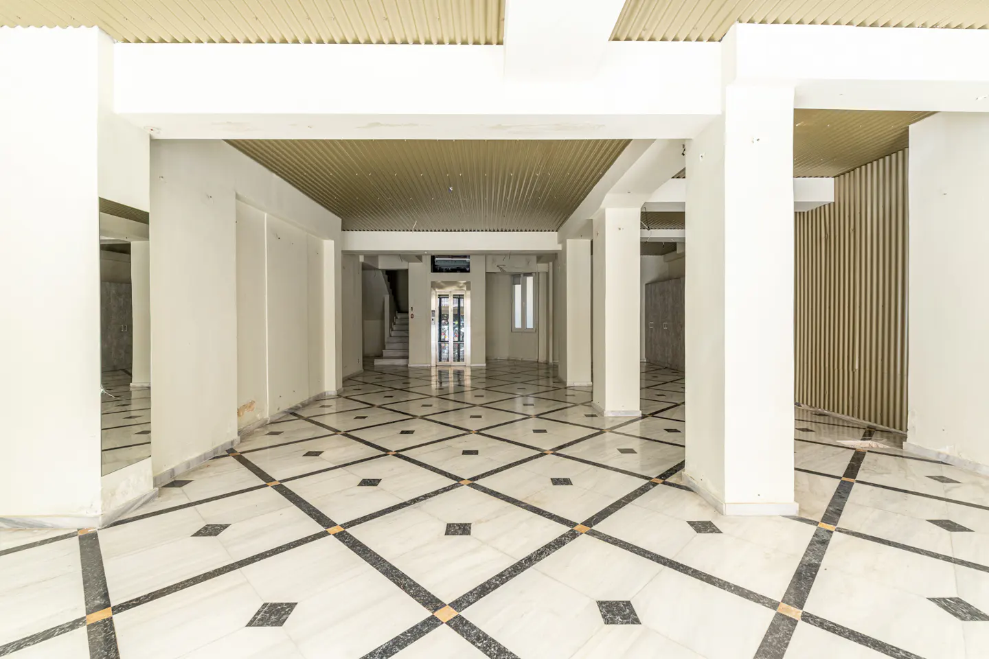 Empty room with white walls, columns, and a black and white diamond-patterned marble floor. A staircase and elevator are visible in the background.