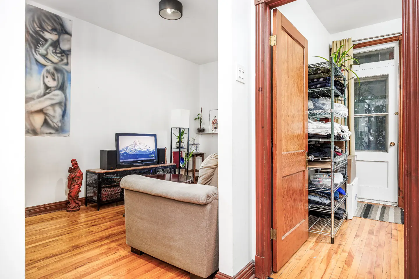 A view of a living room with a TV, couch, and a doorway leading to a hallway with a shoe rack and a door.