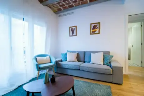 Living room with blue chair, gray sofa with pillows, and oval coffee table on a blue rug. White walls with framed art. Brick ceiling.