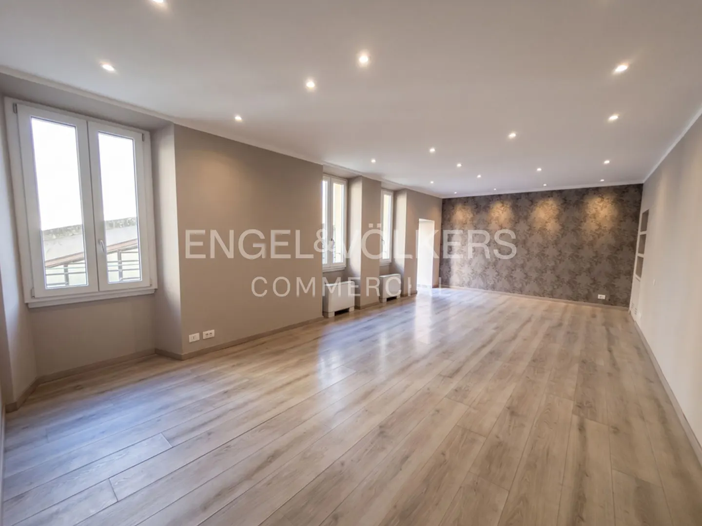 Spacious, empty room with light wood floors, beige walls, recessed lighting, and a dark patterned accent wall.