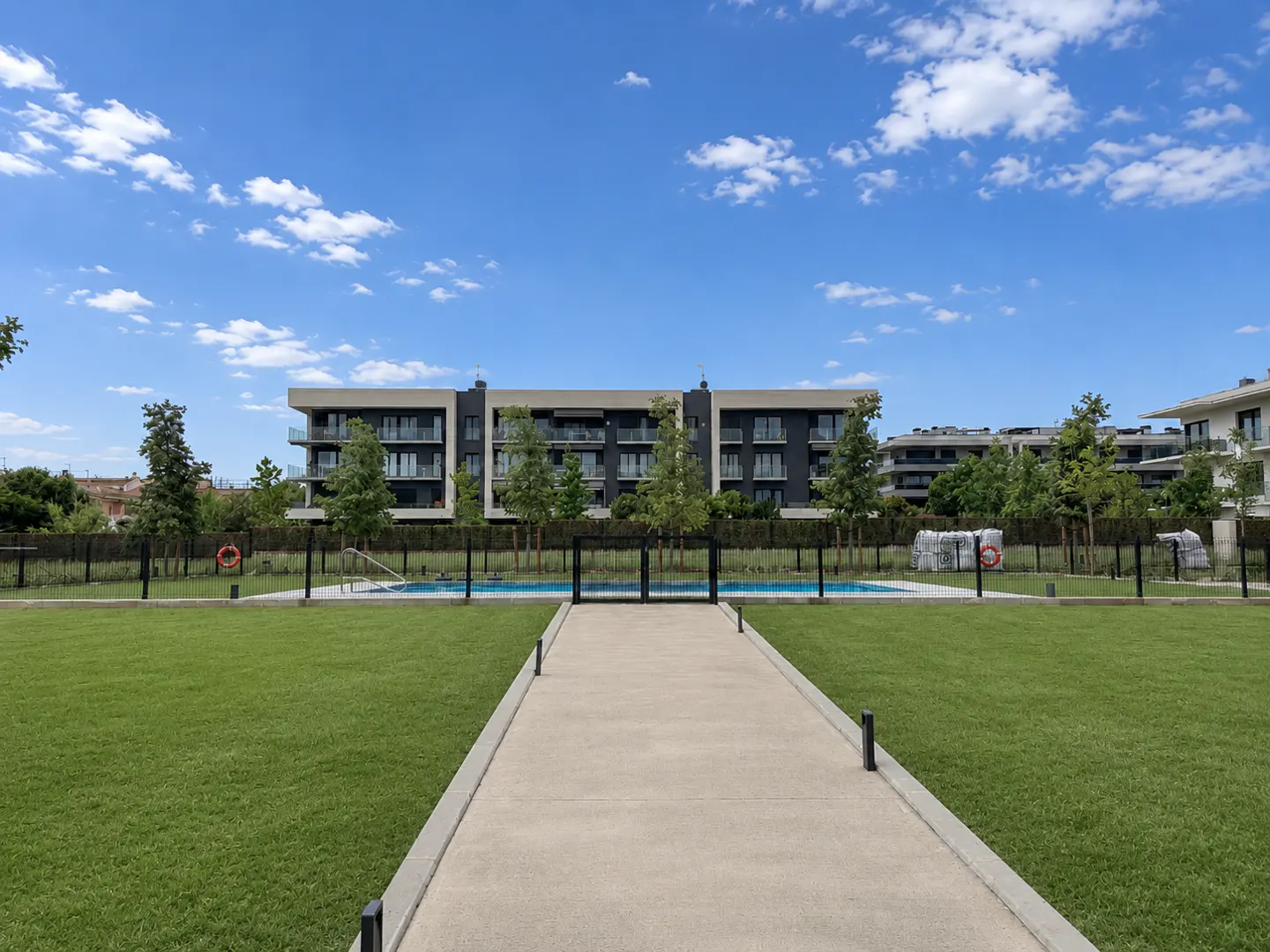 A concrete path leads through a green lawn toward a swimming pool and modern apartment buildings under a blue sky.