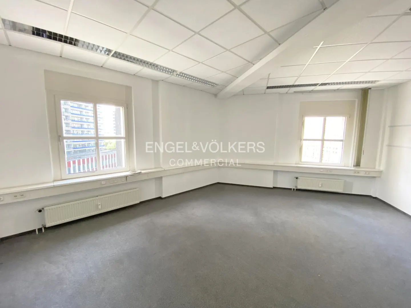 Empty office space with gray carpet, white walls, and two windows. Radiators are under the windows. Engel & Völkers Commercial logo is visible.