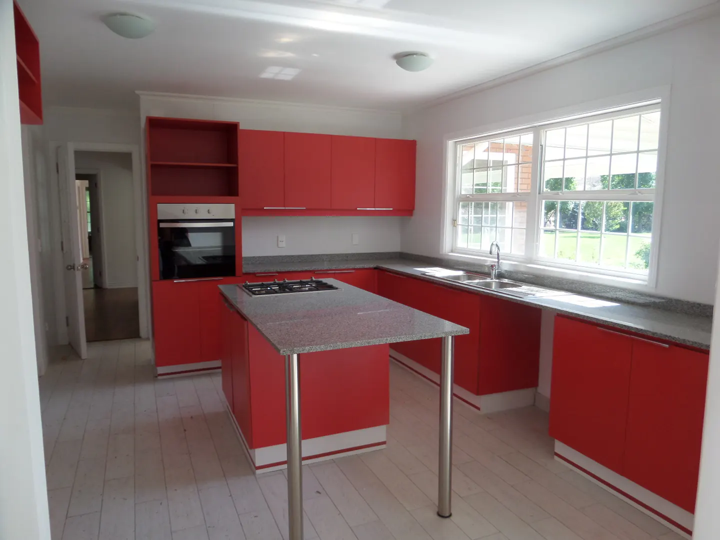 Bright kitchen with red cabinets, gray countertops, and a central island with a cooktop. A window overlooks a green lawn.