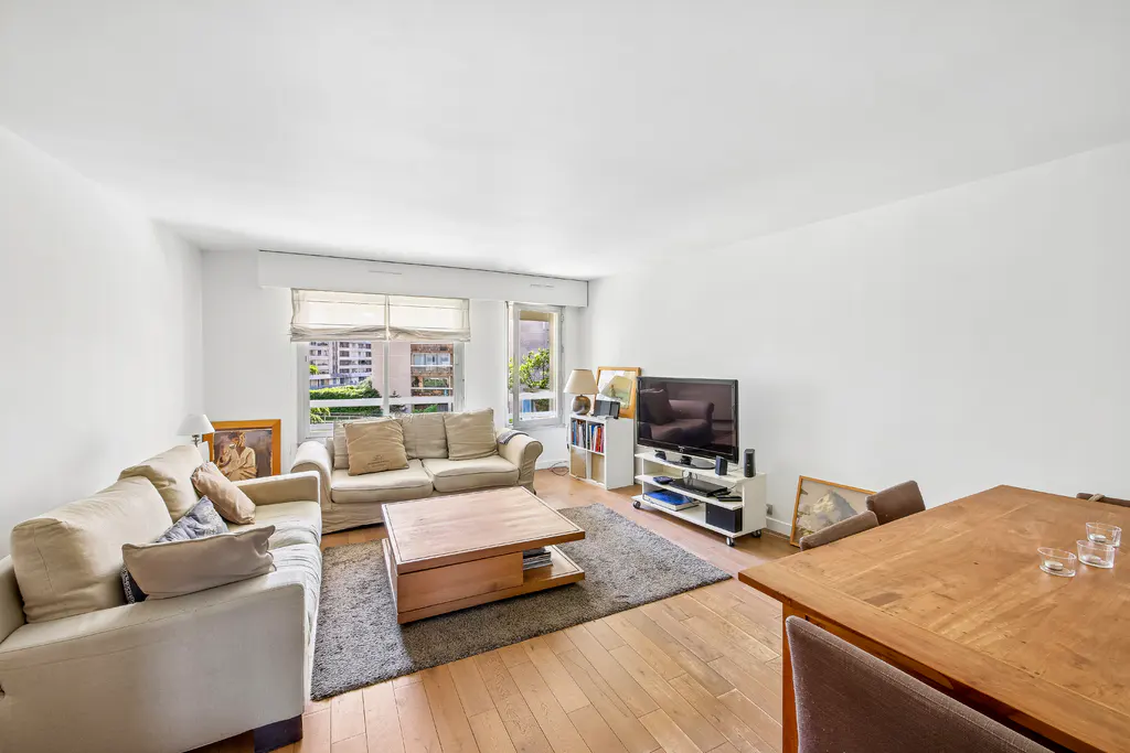 Bright living room with hardwood floors, white walls, and two beige sofas. A wooden coffee table sits on a gray rug. A TV and dining table are also visible.